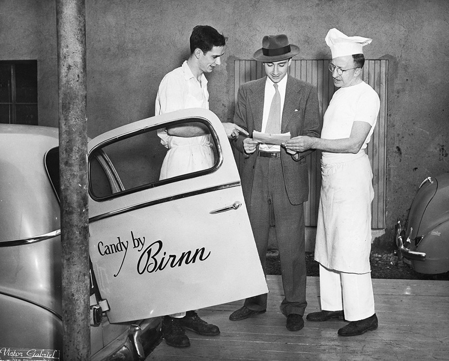 Black and white photo of three men: two are in white candy-maker uniforms and another man is dresssed in a suit reviewing a slip of paper while standing next to a Birnn delivery truck.
