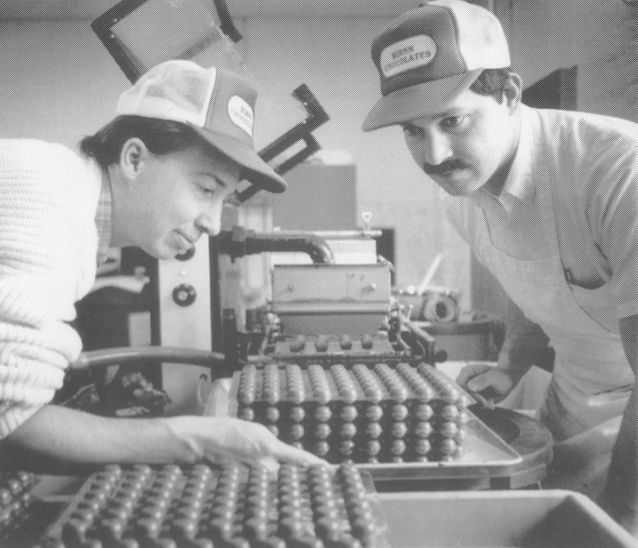 Two men in caps inspecting chocolate truffles on a candy production line.